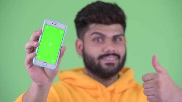 Face of Happy Young Overweight Bearded Indian Man Showing Phone and Giving Thumbs Up