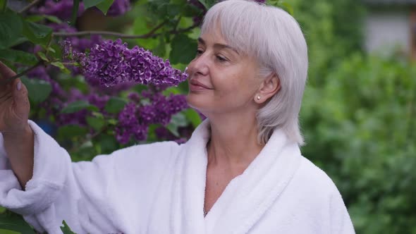 Cheerful Middle Aged Woman Smelling Purple Blossom on Tree in Garden Smiling