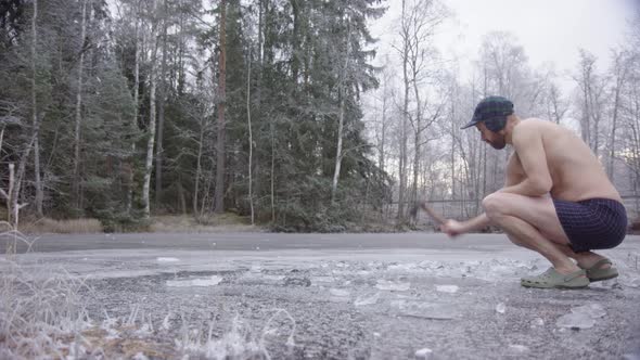 A hipster ice bather enters frame and begins axing the ice to carve a hole