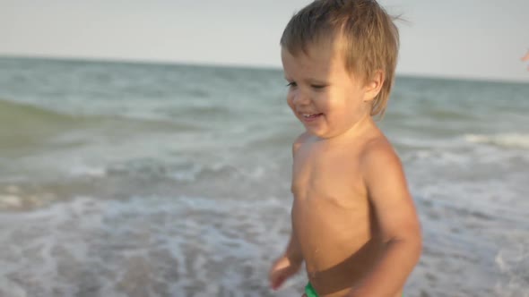 A Girl in a Swimsuit Runs with a Toddler on the Beach with Shells on the Background of the Sea with