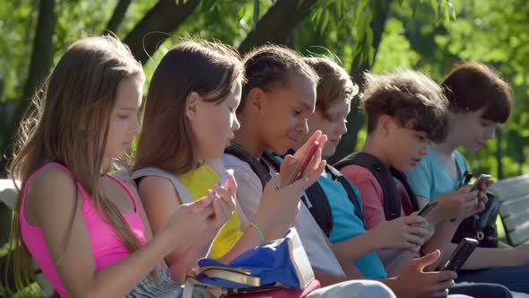 Side View of Diverse Teenager Children Using Cellphone Sitting on Bench in Park