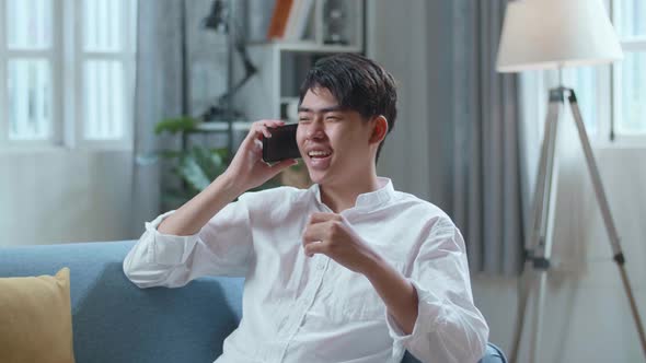 Smiling Asian Man Answering The Call On Smartphone While Lying On Sofa In The Living Room