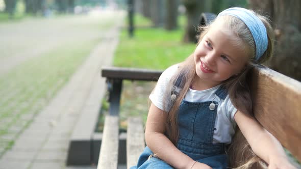 Little Happy Child Girl Sitting on a Bench Smiling Happily in Summer Park