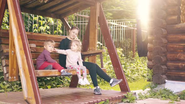 Travelling Young Pretty Mother with Children Swinging on a Wooden Swing Near the Summer House 3