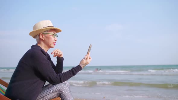 Asian elderly woman chatting with friends on video call while relaxing at the beach.