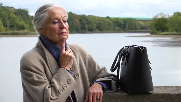 An Elderly Woman Looks Sad, Closeup, a Lake in the Blurry Background