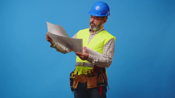 Senior Man Builder in Hardhat Looking at Blueprints Against Blue Background