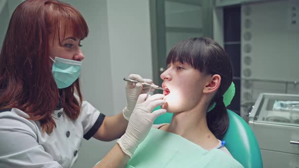 An experienced dentist in a white robe and mask examines the girl's teeth using a dental mirror