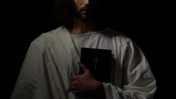 Monk in White Robe Holding Holy Bible Against Dark Background, Christianity