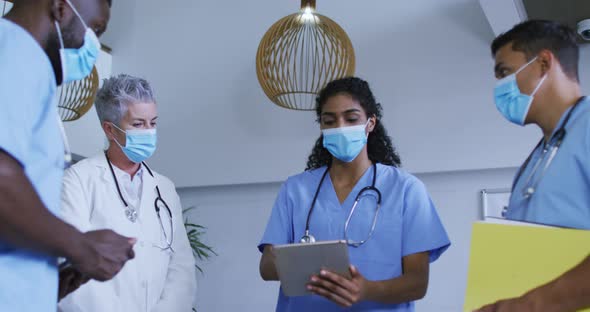Diverse group of male and female doctors in face masks using tablet and discussing in hospital