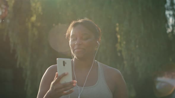 Plus sized African-American woman walking and using mobile phone with earphones in the park in a sum