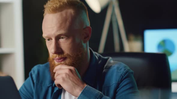 Cheerful Man Speaking on Laptop at Home. Redhead Hipster Working in Office