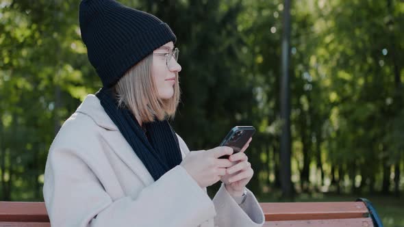 Young Girl Sitting on a Park Bench Looking at Phone and Smiling in Autumn