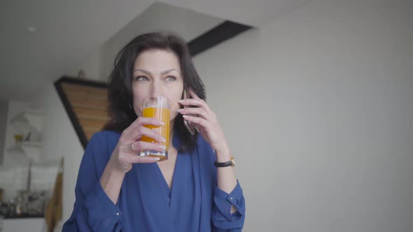 Close-up Portrait of Busy Confident Caucasian Woman Talking on the Phone and Holding Glass of Orange