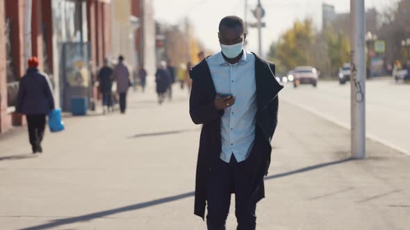 Portrait of African American Business Man in Mask, Smartphone in Hands. City Walk Down the Street