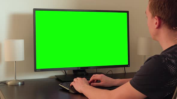 A Man Sits at a Table and Works on a Computer with a Green Screen