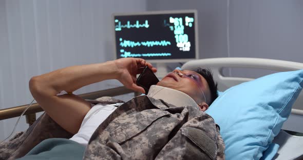 Injured Asian Soldier with Neck Collar Lying in Hospital Bed and Talking on Smartphone