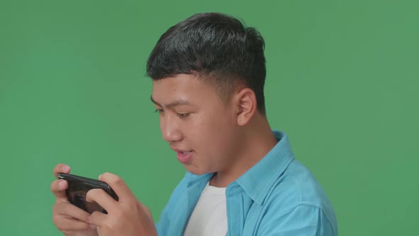 Side View Of Excited Young Asian Boy Playing Video Game With Mobile Phone In Green Screen Studio alt