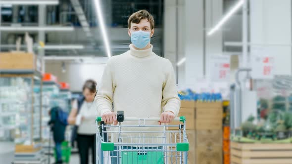 A Man in a Medical Mask Stands in a Supermarket with a Food Cart Timelaps