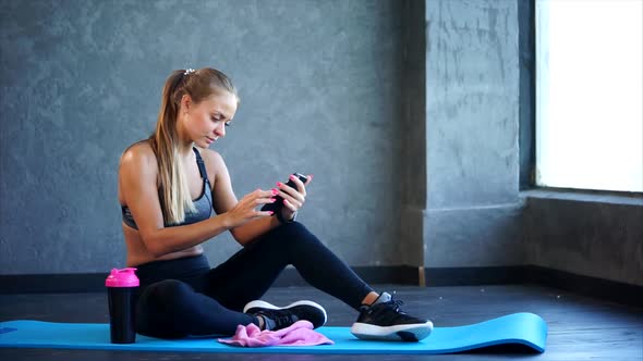 Woman in the Gym with Smartphone. She Sitting on the Mat and Using Smartphone