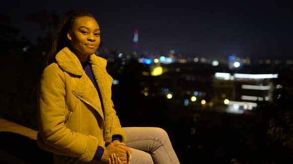 A Young Black Woman Applauds to the Camera with a Smile As She Sits in an Urban Area at Night