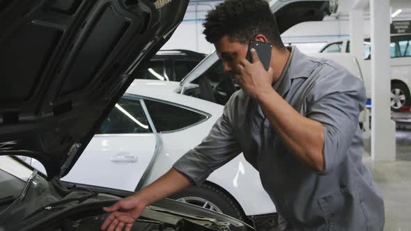 African American male car mechanic looking at an open car engine and talking on a smartphone