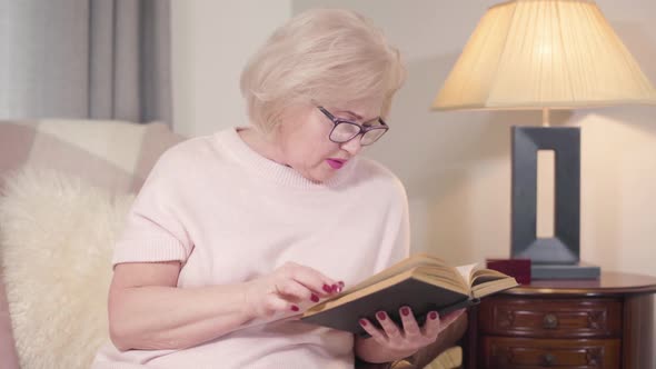 Portrait of Senior Caucasian Blond Woman in Eyeglasses Reading Book at Home. Mature Smart Woman