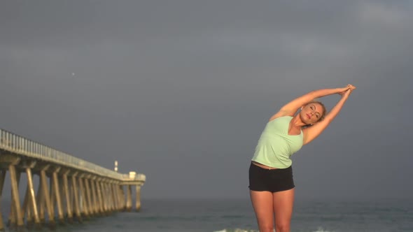 A young woman does yoga on the beach next to a pier.