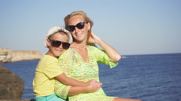 Cute Baby Kisses His Mother on the Cheek, He Is Sitting on the Beach on a Sunny Clear Day. Vacation