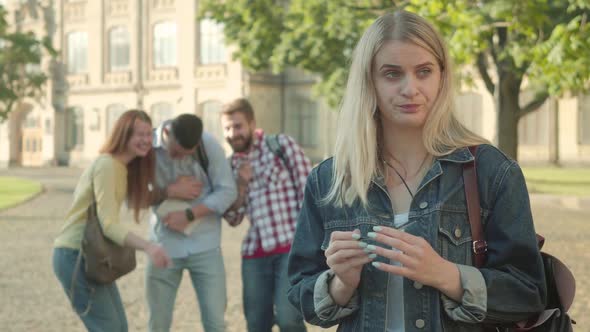 Beautiful Shy Blond Girl Smiling at Camera As People Laughing at Her at the Background. Confident