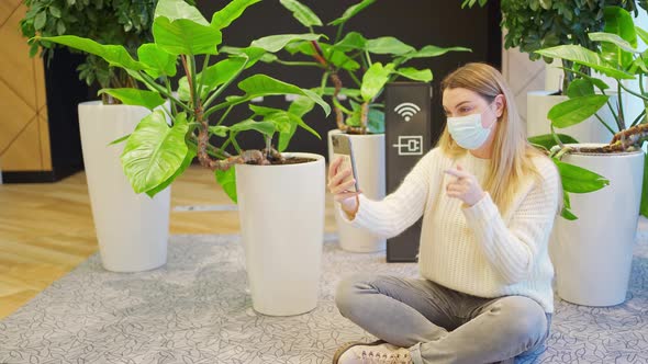 a Young Woman in a Protective Mask Sits on the Floor Near Home Green Plants and a Wifi Sign and