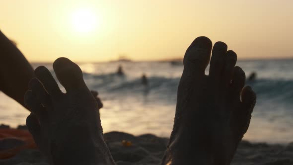 POV Silhouette of Feet of Young Man Lying on Sandy Beach By Ocean During Sunset