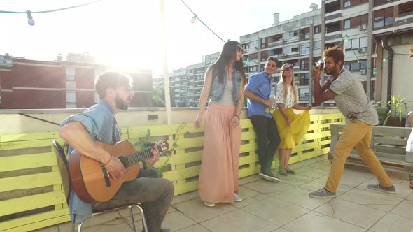 Friends having fun on rooftop terrace, playing guitar, dancing and taking photos