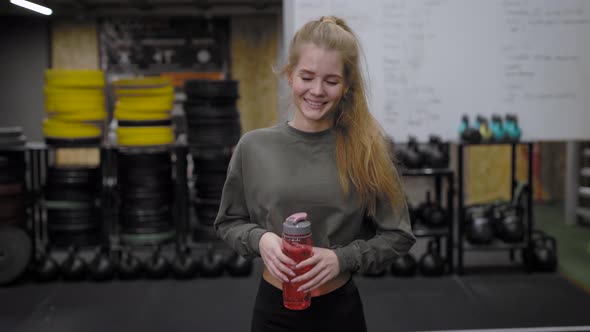 Happy Smiling Girl Is Walking Across Gymnastic Hall Before Training, Holding Bottle with Water