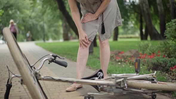 Unrecognizable Middle Aged Caucasian Woman Rubbing Injured Knee with Bike Lying at Front on Summer