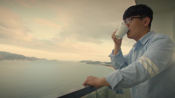 Handsome Dreamy Asian Middle Aged Man Stands in the Balcony with Great Views and Drinks Hot Coffee