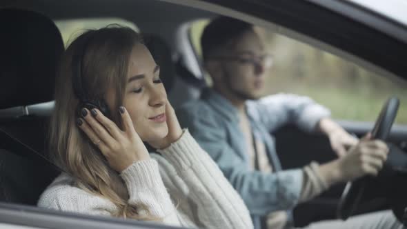 Side View of Happy Relaxed Young Woman in Headphones Dancing with Blurred Man Sitting on Driver's