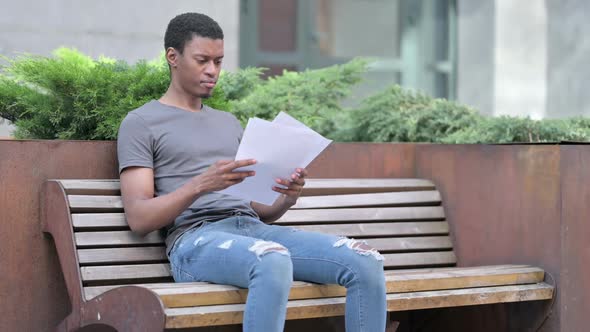 Young African Man Reading Documents Sitting on Bench
