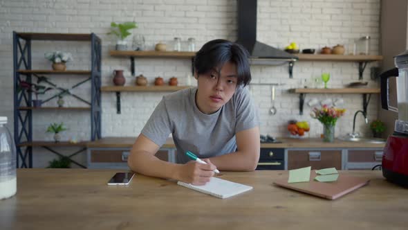 Wide Shot of Young Man Writing in Slow Motion and Looking at Camera Sitting at Kitchen Table