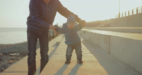 Dad Leads the Little Son By the Arms Teaches Him to Walk