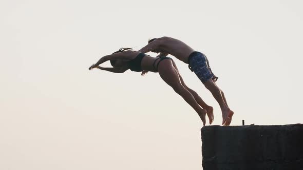 Young Woman and Man Synchronously Doing Frontflip From a Pier Into the Sea During Beautiful Sunrise