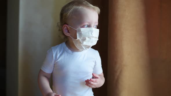 baby in a protective medical mask looks out the window from behind a curtain alt