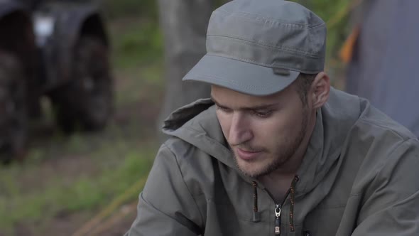 Young Adult Male Tourist Sits on Log Near Fire in Forest. Human Looks at Forest Fire on Nature. Man