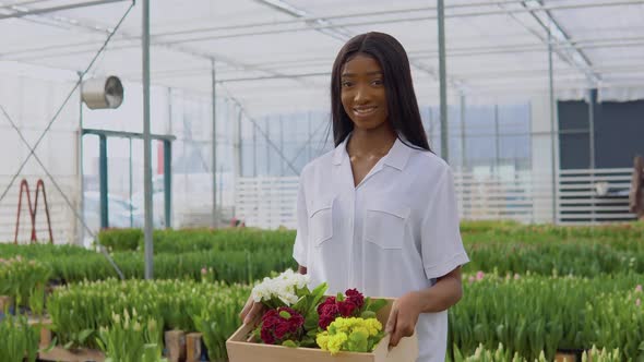Beautiful Young African American Girl in a White Shirt Stands in a Greenhouse and Holds a Craft