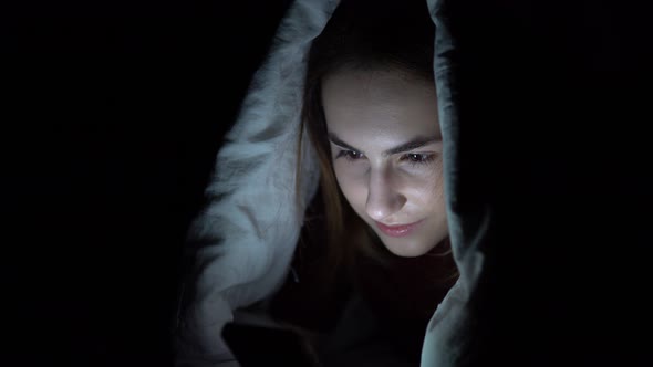 A Young Woman Lies Under a Blanket with a Phone in Her Hands. A Woman in the Dark Uses a Pad. Modern