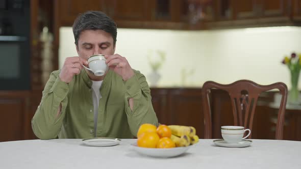 Potrait of Sad Lonely Caucasian Man Drinking Herbal Tea Looking at Empty Chair and Cup on Table