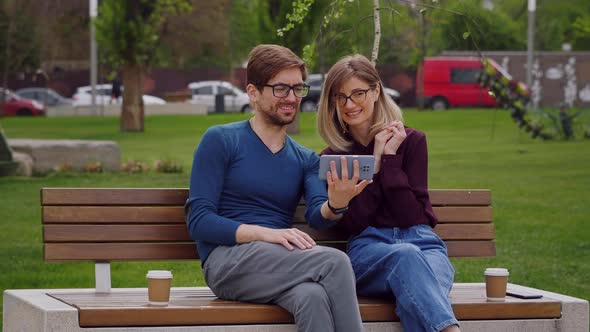 Man And Woman Looking smiling At Phone outdoor sitting on a bench.