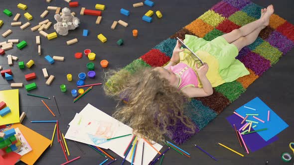 Long-Haired Little Girl Lying on Colorful Carpet and Playing Games on Tablet