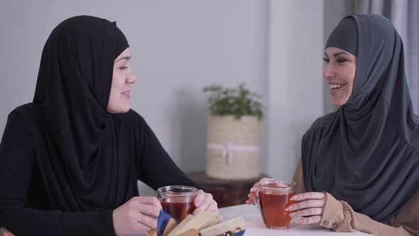 Close-up of Two Happy Muslim Women Talking and Laughing Indoors. Female Friends Sitting at the Table