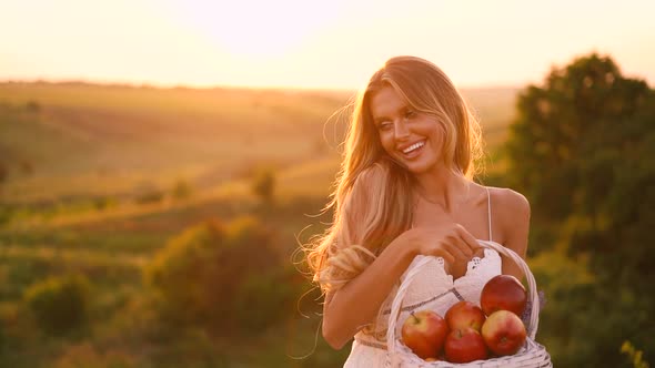 Beautiful sexy blonde girl in white dress posing in a field at sunset with a basket of fruit	